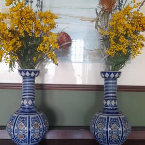 May include: Two blue and white ceramic vases with intricate patterns hold yellow flowers and dried plants. The vases have a bulbous shape and a long neck. The background features a painting with a landscape scene.