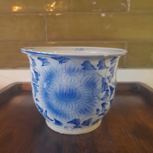 May include: A white ceramic planter with a blue and white floral design. The planter has a wide mouth and a rounded bottom.