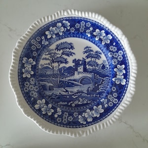 May include: A round ceramic plate with a white, ruffled edge and a blue and white landscape design. The central image depicts a bridge, trees, and a castle. Floral patterns adorn the plate's rim.