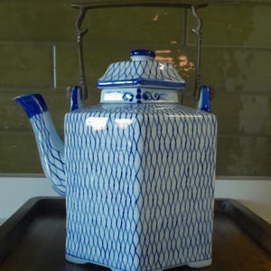 May include: A white and blue ceramic teapot with a square body and a pagoda-style lid. The teapot features a blue lattice pattern and a metal handle. The spout and handle are blue. The teapot sits on a wooden tray.