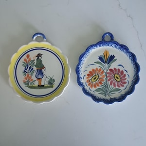 May include: Two decorative ceramic plates with scalloped edges. One plate has a yellow border and a figure with a watering can. The other plate has a blue border and floral designs in orange, red, and blue. Both plates have a loop for hanging.