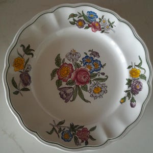 May include: White ceramic plate with a scalloped edge and a green trim. The plate features a floral design with pink, yellow, blue, and purple flowers. The flowers are arranged in a bouquet in the centre and along the rim.