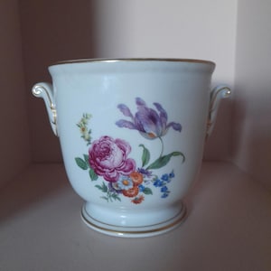 Könnte beinhalten: Weißer Keramik-Blumentopf mit goldenen Verzierungen und Griffen. Der Topf hat ein Blumendesign mit einer großen rosa Rose und einer lila Tulpe. Ein dekorativer Artikel für den Innenbereich.