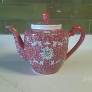 May include: A red ceramic teapot with a white floral design and a matching lid. The teapot has a curved spout and handle, and the lid has a small knob. The teapot is decorated with a repeating pattern of swirls.