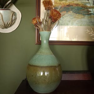 May include: A ceramic vase with a light green and brown glaze holds dried flowers. The vase has a rounded body and a slender neck. The vase is approximately 12 inches tall and sits on a dark wooden surface.