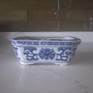 May include: A rectangular ceramic planter with a white base and blue floral patterns. The rim features a geometric cross-hatch design, while the sides display stylized lotus-like flowers and scrolling vines. The vessel stands on four small feet.
