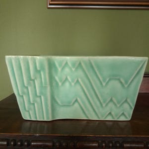 May include: A rectangular ceramic vase in a light mint green colour. The exterior surface features a raised, moulded geometric pattern consisting of vertical stepped lines on the left side and repeating horizontal zigzag lines across the rest.