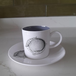 May include: A white ceramic coffee cup with a purple rim and a black and white illustration of an apple on the front. The cup is sitting on a white saucer with a matching apple illustration.