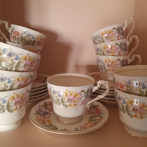 Peut inclure: Un ensemble de tasses et soucoupes blanches avec un motif floral rose, jaune et bleu. Les tasses ont des bordures et des anses dorées. L'ensemble comprend plusieurs tasses et soucoupes, empilées et disposées sur une étagère.
