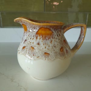 May include: A ceramic jug with a handle and spout. The jug has a cream-coloured base, with a speckled brown and white pattern on the upper portion. The rim and handle are a solid brown colour.