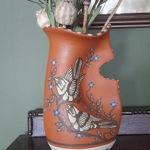 May include: Earthenware vase in a terracotta hue, featuring a bird motif and a distinctive cut-out. The vase is adorned with white and blue floral details. It contains dried seed heads and foliage, forming a rustic decorative arrangement.