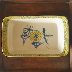 May include: A rectangular ceramic baking dish with a yellow and black border. The dish features a stylised design of green leaves, blue and yellow fish, and a yellow sun.