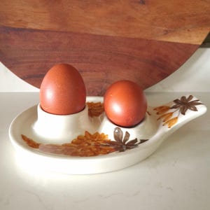 May include: A cream-colored ceramic egg holder with two circular indentations, each holding a brown egg. The tray features a decorative floral pattern in shades of orange and brown, with a handle extending from one side of the oval-shaped base.