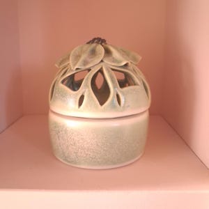 May include: A cylindrical ceramic jar with a light green speckled glaze. The lid features a sculpted flower design with cut-out petal shapes and a dark brown center. The base is a simple, smooth cylinder that matches the color and texture of the lid.