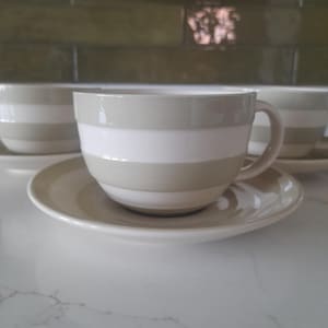 May include: A white ceramic mug with a black handle and the text "But First, Coffee" in a bold, black font. The mug is set against a blurred background, suggesting a cozy setting.