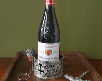 May include: A silver wine bottle holder with a grapevine design, a cork stopper, and a corkscrew. The bottle holder is holding a bottle of red wine labeled "H. Valrasque Châteauneuf-du-Pape".