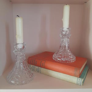 May include: Two clear crystal candle holders with lit white candles sit on a stack of books. The books have orange and green covers. The candle holders have a detailed, cut-glass design.
