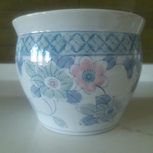 May include: White ceramic planter with a blue and pink floral design. The planter features a blue lattice pattern around the top and is decorated with stylized flowers and leaves. The pot has a slightly flared rim.