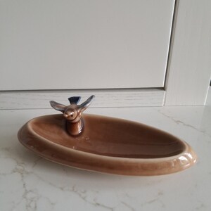 May include: An oval-shaped ceramic trinket dish with a glossy light brown finish. A small, sculpted bird with blue and brown wings is attached to one side of the rim, facing inward toward the centre of the shallow, smooth, concave basin.