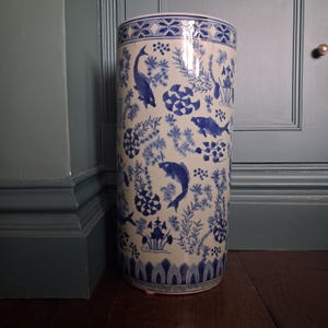 May include: A cylindrical ceramic umbrella stand featuring a white base with a blue hand-painted pattern of swimming fish, aquatic plants, and geometric floral motifs. The top and bottom rims are decorated with a repeating blue border design.
