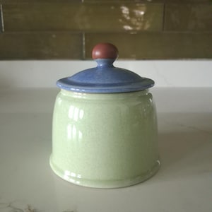 May include: A ceramic canister with a light green speckled body and a smooth, rounded blue lid. The lid features a small, dark reddish-brown spherical knob at the top. The container has a cylindrical shape with a slightly flared base and a glossy finish.