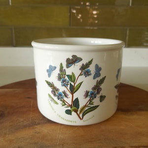 May include: A white ceramic jar with a botanical design featuring blue flowers, green leaves, and butterflies. The jar has a cylindrical shape and is decorated with the words "Veronica Chamaedrys" and "Speedwell".