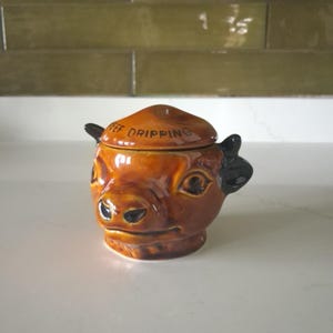 May include: A brown ceramic pot shaped like a cow head with a matching lid. The lid features the text BEEF DRIPPING in black lettering. The pot has dark brown ears and a glossy finish, designed to hold beef dripping or other kitchen fats.
