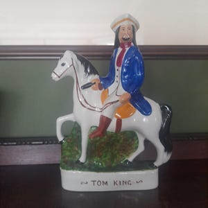 May include: A ceramic figurine of a man on horseback, painted in white, blue, and red. The man wears a tricorn hat and a blue coat. The horse is white with black accents. The base reads "TOM KING".