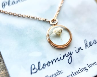 May include: A gold necklace with a delicate chain and a round pendant featuring a preserved baby's breath flower. The pendant has a clear glass dome and a gold rim. The card reads "Planted on Earth, Blooming in Heaven, Baby's Breath: A symbol of everlasting love."