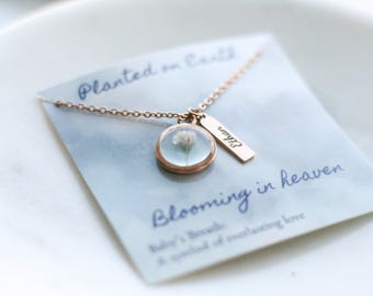 May include: A gold necklace with a small round pendant containing a pressed white flower. The pendant is attached to a small gold tag with the word "Ethan" engraved on it. The necklace is on a blue card with the text "Planted on Earth, Blooming in Heaven, Baby's Breath, A symbol of everlasting love."