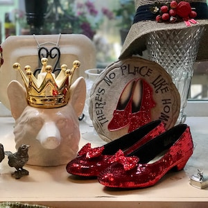 May include: A white ceramic dog figurine wearing a gold crown, a glass vase with a gold crown and red jewels, a red and gold sign that says "There's No Place Like Home", and two red sequined shoes with bows.