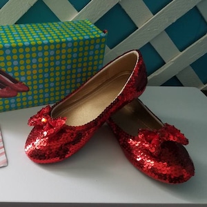 May include: A pair of red sequined shoes with a bow on each toe. The shoes are sitting on a white surface next to a gift box wrapped in green and blue polka dot paper and a pink and white striped card with the text "Thank you!" written on it.