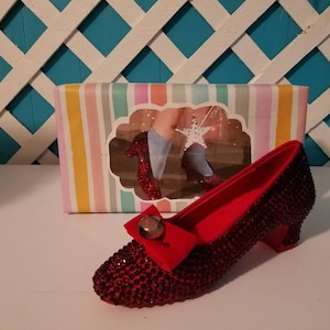 May include: A pair of red ruby slipper shoes with a sparkly finish and a red bow. The shoes are on a white surface with a box behind them. The box has a colorful striped design and a picture of a person wearing the shoes.