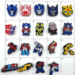 May include: A collection of 20 colorful, cartoonish character charms, including robot faces and figures. The charms are in various colors, including blue, red, yellow, and black. The collection is arranged in a grid.