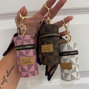 May include: Three cylindrical keychains in pink, brown, and white, each with a checkered pattern and a gold zipper. The keychains have gold hardware and a small leather tag that reads "Vogue New Famous."