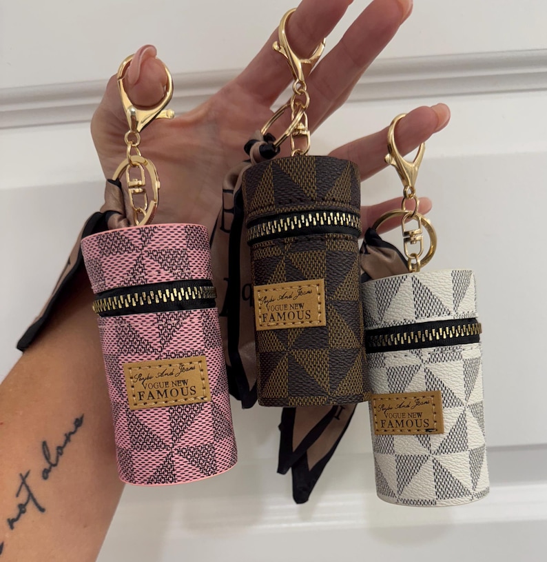 May include: Three cylindrical keychains in pink, brown, and white, each with a checkered pattern and a gold zipper. The keychains have gold hardware and a small leather tag that reads "Vogue New Famous."