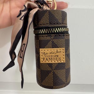 May include: Brown and tan checkered cylindrical keychain case with a gold zipper and a gold keyring. A small tan and black scarf is attached. The case has a small rectangular label that reads "Saybe And Jeans VOGUE NEW FAMOUS."