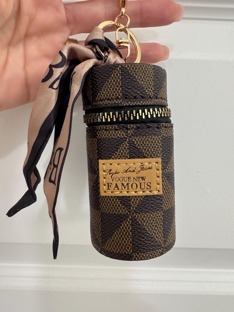 May include: Brown and tan checkered cylindrical keychain case with a gold zipper and a gold keyring. A small tan and black scarf is attached. The case has a small rectangular label that reads "Saybe And Jeans VOGUE NEW FAMOUS."