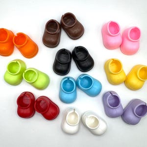 May include: A collection of colorful doll shoes in various shades, including orange, brown, black, pink, yellow, green, red, blue, white, and purple. The shoes are small, with a rounded toe and a circular opening.