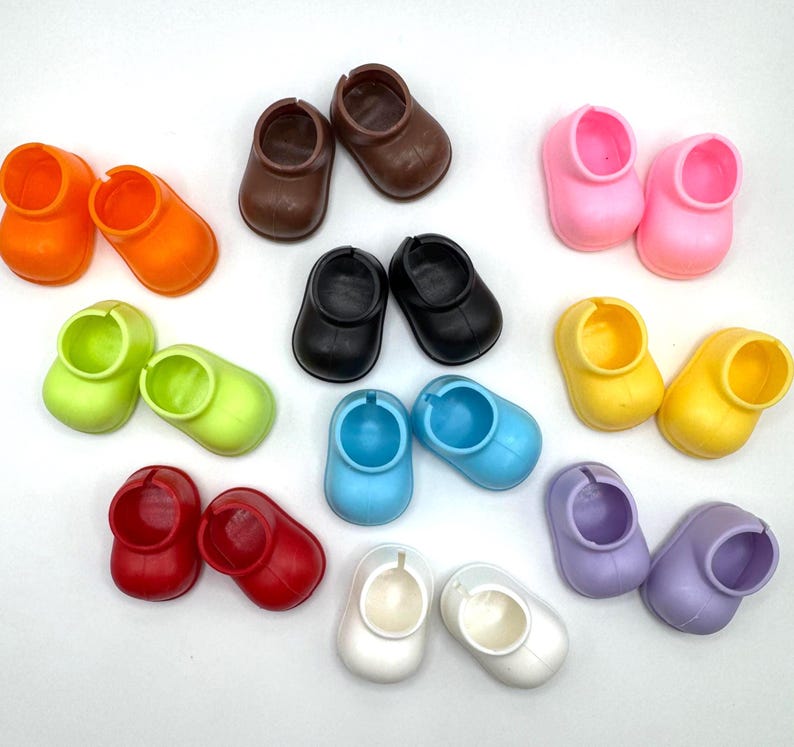 May include: A collection of colorful doll shoes in various shades, including orange, brown, black, pink, yellow, green, red, blue, white, and purple. The shoes are small, with a rounded toe and a circular opening.