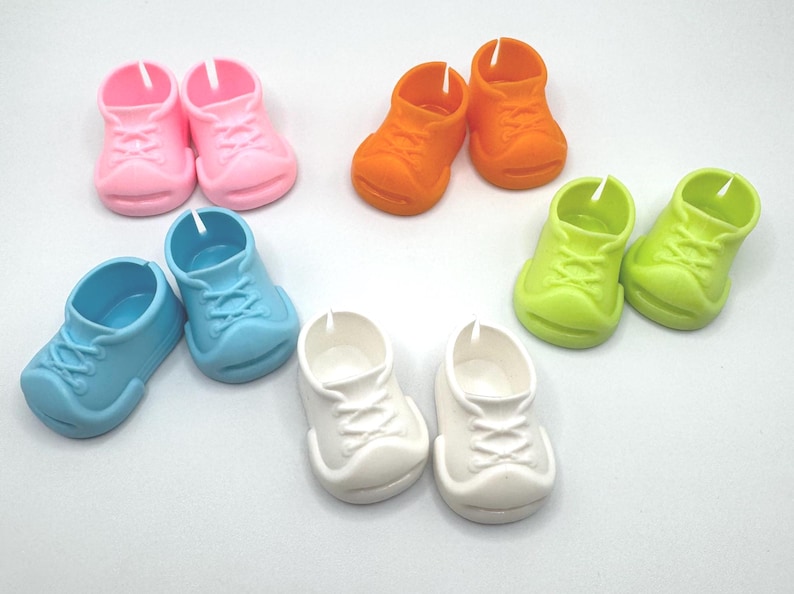 May include: A collection of colorful miniature toy sneakers in pink, orange, light blue, green, and white. The shoes are arranged in pairs, showcasing their small size and playful design. These plastic shoes are likely for dolls or other toys.