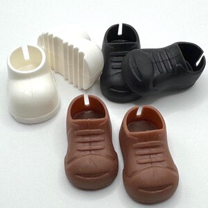 May include: A collection of miniature toy shoes in various colors. There are two brown shoes, two black shoes, and one white shoe. A white sole is also present. The shoes have a classic lace-up design.