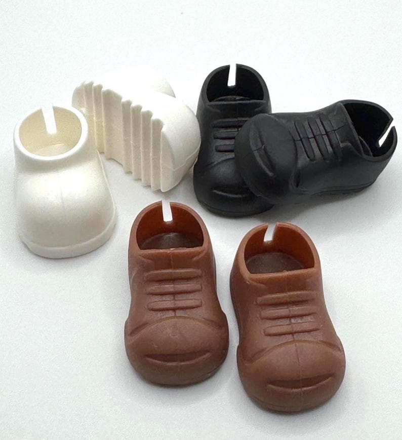 May include: A collection of miniature toy shoes in various colors. There are two brown shoes, two black shoes, and one white shoe. A white sole is also present. The shoes have a classic lace-up design.
