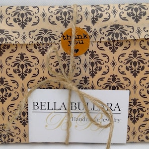 May include: A rectangular package wrapped in tan paper with a dark brown damask pattern, tied with natural jute twine. A round orange sticker reads thank you. A white rectangular card features the black text BELLA BULLARA Handmade Jewelry.