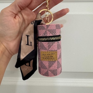 May include: Pink and black checkered cylindrical keychain case with a gold zipper and a gold keychain ring. The case has a small gold label that reads "Vogue New Famous". A small scarf with the letter "L" is attached.
