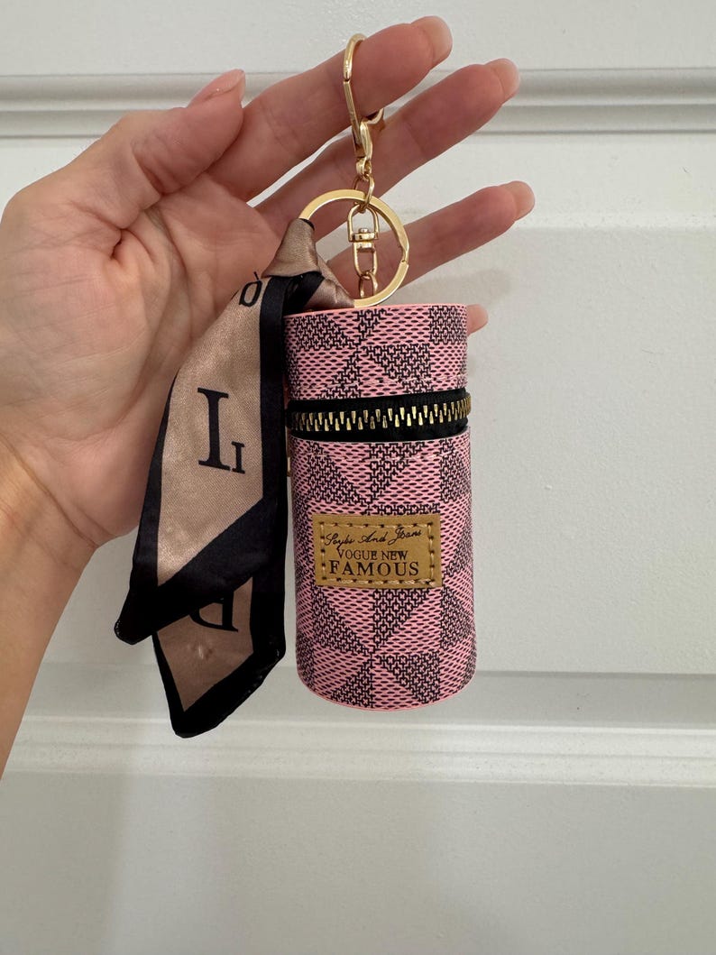 May include: Pink and black checkered cylindrical keychain case with a gold zipper and a gold keychain ring. The case has a small gold label that reads "Vogue New Famous". A small scarf with the letter "L" is attached.