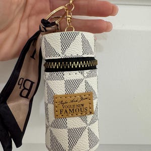 May include: A white and black geometric pattern keychain pouch with a gold zipper and hardware. The pouch has a small brown leather tag with the words "Vogue New Famous" printed on it. A black and tan scarf is attached.