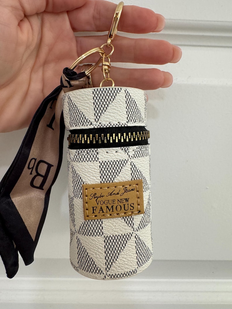 May include: A white and black geometric pattern keychain pouch with a gold zipper and hardware. The pouch has a small brown leather tag with the words "Vogue New Famous" printed on it. A black and tan scarf is attached.