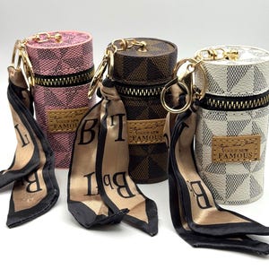May include: Three cylindrical keychains in pink, brown, and white, each with a zipper closure and gold-tone hardware. The keychains feature a checkered pattern and are adorned with a silk scarf with black lettering. The label reads "Vogue New Famous."