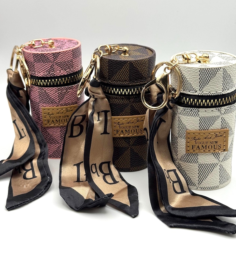 May include: Three cylindrical keychains in pink, brown, and white, each with a zipper closure and gold-tone hardware. The keychains feature a checkered pattern and are adorned with a silk scarf with black lettering. The label reads "Vogue New Famous."